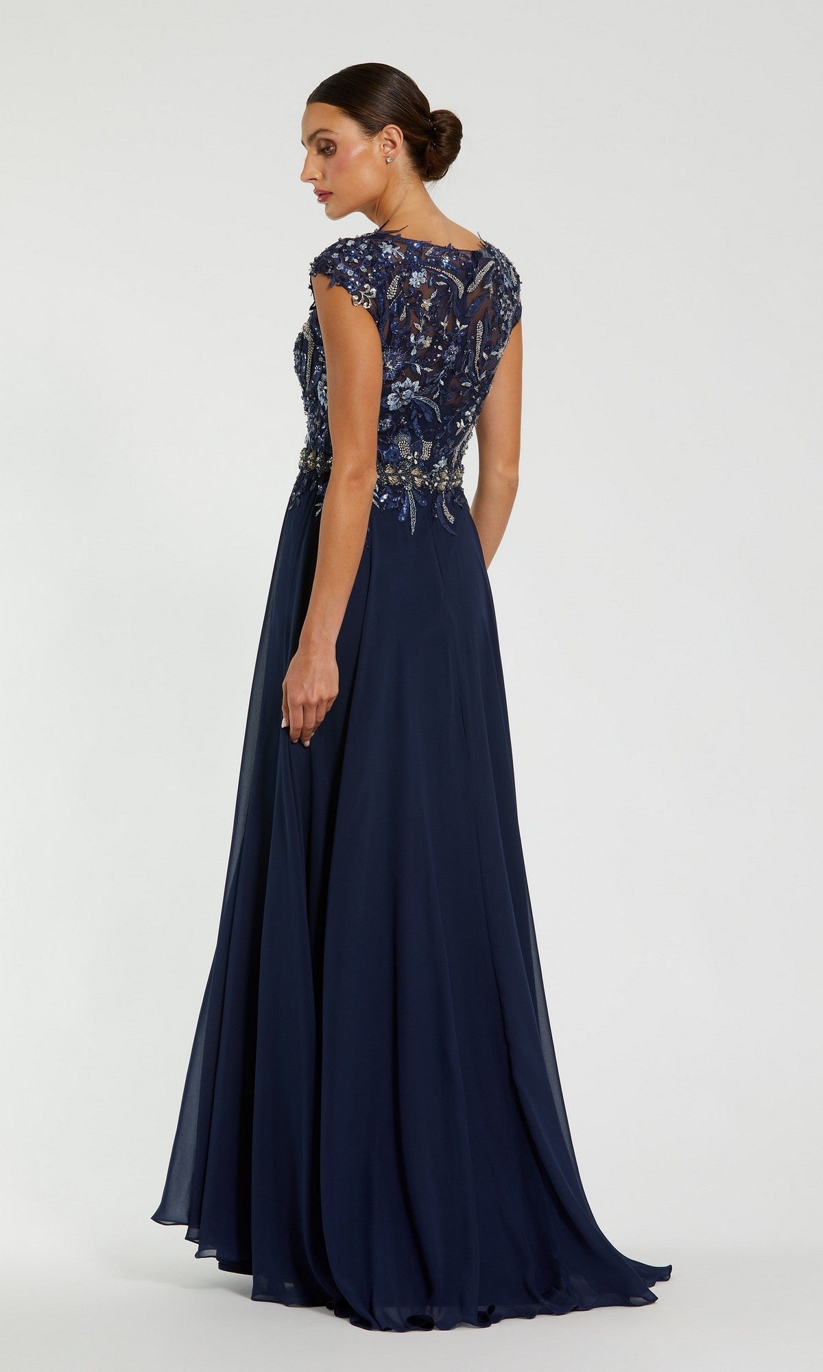 A woman wearing the Mac Duggal 79365 Cap-Sleeve Long Formal Dress in navy blue, featuring an embellished bodice, stands facing away and looks over her shoulder against a plain background.